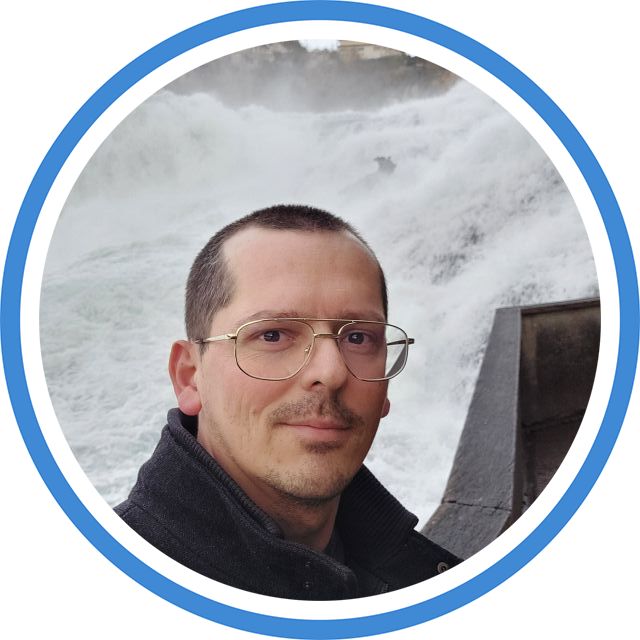 Rundes Porträtfoto von Christian Schöniger. Ein Mann mit Brille und dunkler Jacke steht in der Natur vor einem fließenden Gewässer.